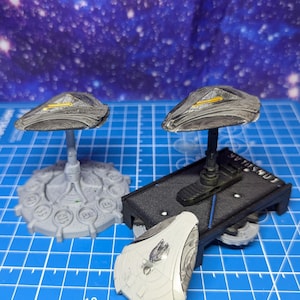 May include: Two grey and silver model spaceships with detailed designs on a blue grid surface. One spaceship is on a black base with the word "LUMINOUS" printed on it. The other spaceship is on a grey base with a circular design.