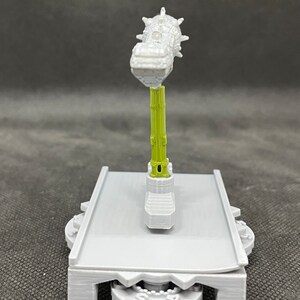 May include: A 3D printed model of a grey and green mechanical device with a spiked top. The device is mounted on a grey base with a gear system.
