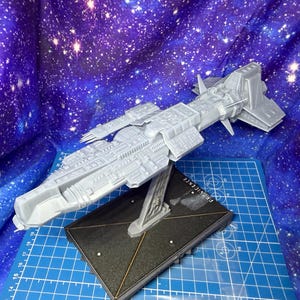 Puede incluir: Modelo de nave espacial gris con detalles intrincados, montado sobre una base negra. La base tiene la palabra "PERSISTENCE" impresa. El modelo está situado contra un fondo de una galaxia estrellada.