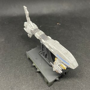 May include: A gray and black spaceship model with a blue stripe on the side, mounted on a black stand. The spaceship has a long, thin body and a pointed nose.