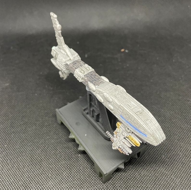 May include: A gray and black spaceship model with a blue stripe on the side, mounted on a black stand. The spaceship has a long, thin body and a pointed nose.