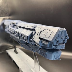 May include: A detailed grey model spaceship, showcasing intricate mechanical designs. The model is mounted on a clear stand, with a dark background. The spaceship features multiple compartments, vents, and weapon ports.