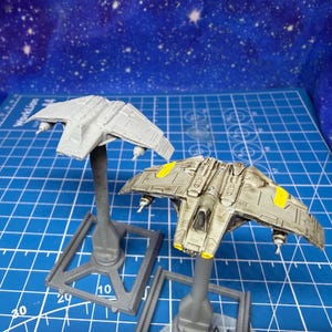 May include: Two miniature model starfighters, one unpainted and the other painted in shades of gray with yellow accents. The models are mounted on gray stands against a blue background with a grid pattern and a starry sky.