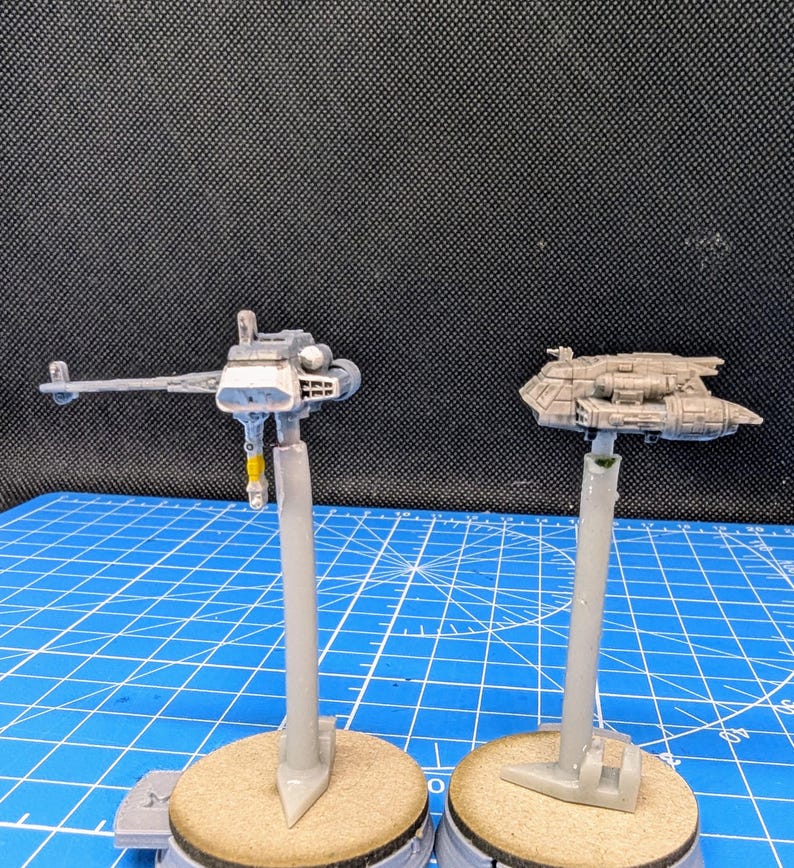 Op de afbeelding: Twee ongeverfde miniatuur-ruimteschepen op displaystandaards. Het ruimteschip links is een grijze en zilveren jager met een lange, dunne vleugelspanwijdte. Het ruimteschip rechts is een grijs en bruin transport met een afgeronde vorm. Beide ruimteschepen staan op grijze plastic standaards.
