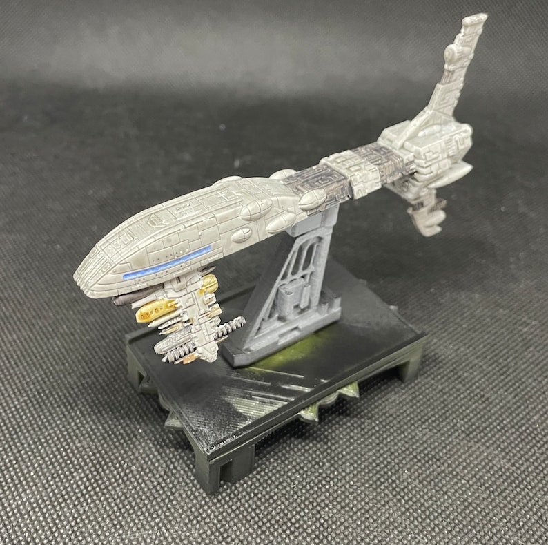 May include: A gray and white spaceship model with a detailed design, mounted on a black base with a textured surface. The spaceship has a long, narrow body with a pointed nose and a large, round engine at the back. The model is painted in a realistic style, with a variety of shades of gray and white.