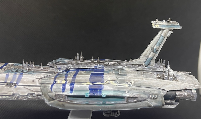 Puede incluir: Un modelo detallado de una gran nave espacial gris con detalles azules. La nave espacial tiene un cuerpo largo y estrecho con una gran cubierta plana en la parte superior. Hay varias cubiertas y estructuras m&aacute;s peque&ntilde;as a lo largo de los lados de la nave. La nave tiene una gran ventana rectangular en el lateral y una torre alta y delgada en la parte posterior.