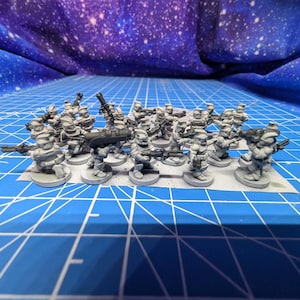 May include: A collection of miniature gray figurines, each depicting a soldier with a weapon. The figurines are arranged on a blue grid surface, with a starry backdrop. The miniatures are likely for tabletop gaming or model building.
