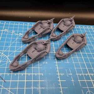 May include: Four gray, futuristic-looking components are arranged on a blue grid cutting mat. Each component has a circular central element with a raised, detailed surface, connected to a curved handle-like structure. The components appear to be 3D-printed.