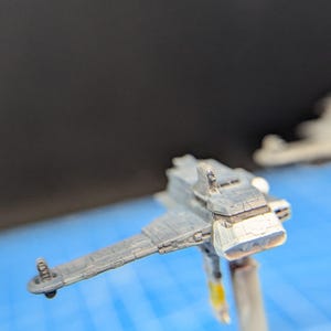 Op de afbeelding: Een grijs en wit miniatuurmodel van een ruimteschip met een gedetailleerd ontwerp. Het model is gemonteerd op een transparante plastic standaard.