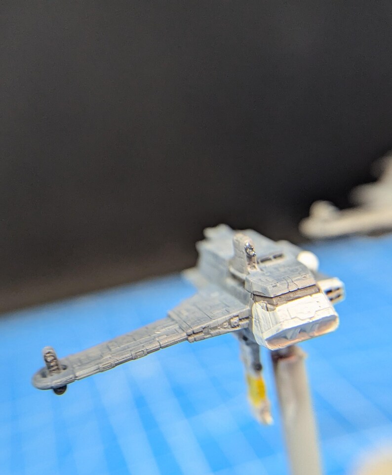 Op de afbeelding: Een grijs en wit miniatuurmodel van een ruimteschip met een gedetailleerd ontwerp. Het model is gemonteerd op een transparante plastic standaard.