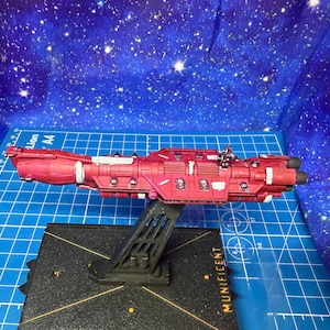May include: A red spaceship model with white detailing, mounted on a black stand. The model features intricate designs and is set against a blue backdrop with a starry pattern. The word "MUNIFICENT" is visible on the base.