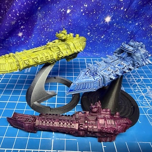 Könnte beinhalten: Drei Miniatur-Raumschiffmodelle in Gelb, Blau und Lila. Das gelbe Schiff steht auf einem silbernen Ständer, das blaue Schiff auf einem schwarzen Ständer und das lila Schiff auf einem blauen Raster. Der Hintergrund ist ein Sternenhimmel.