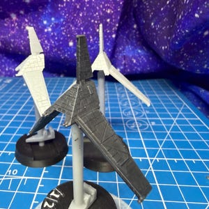 May include: Three miniature spaceship models. One is dark gray, and two are white. The models are mounted on stands with black bases. The background is a blue surface with a grid pattern and a starry design.