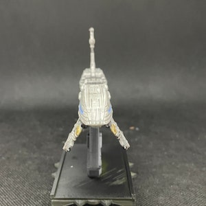 May include: A silver spaceship model with blue accents and a black base. The spaceship has a long, thin body with a pointed nose and two wings. The base is rectangular and has a textured surface.