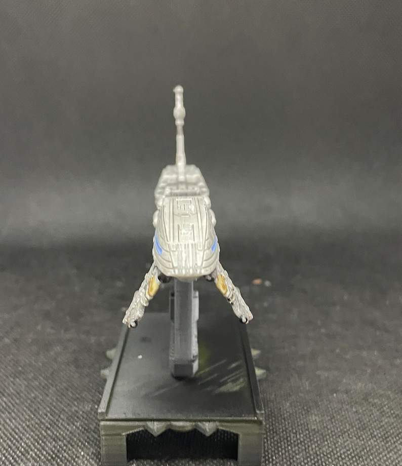 May include: A silver spaceship model with blue accents and a black base. The spaceship has a long, thin body with a pointed nose and two wings. The base is rectangular and has a textured surface.