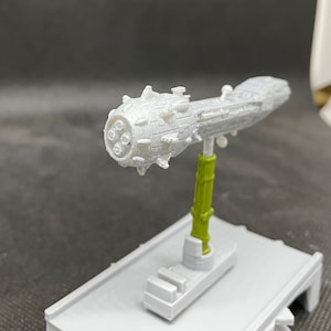 May include: A white 3D printed model of a spaceship with a spiky, round front section and a long, thin body. The model is mounted on a green stand.