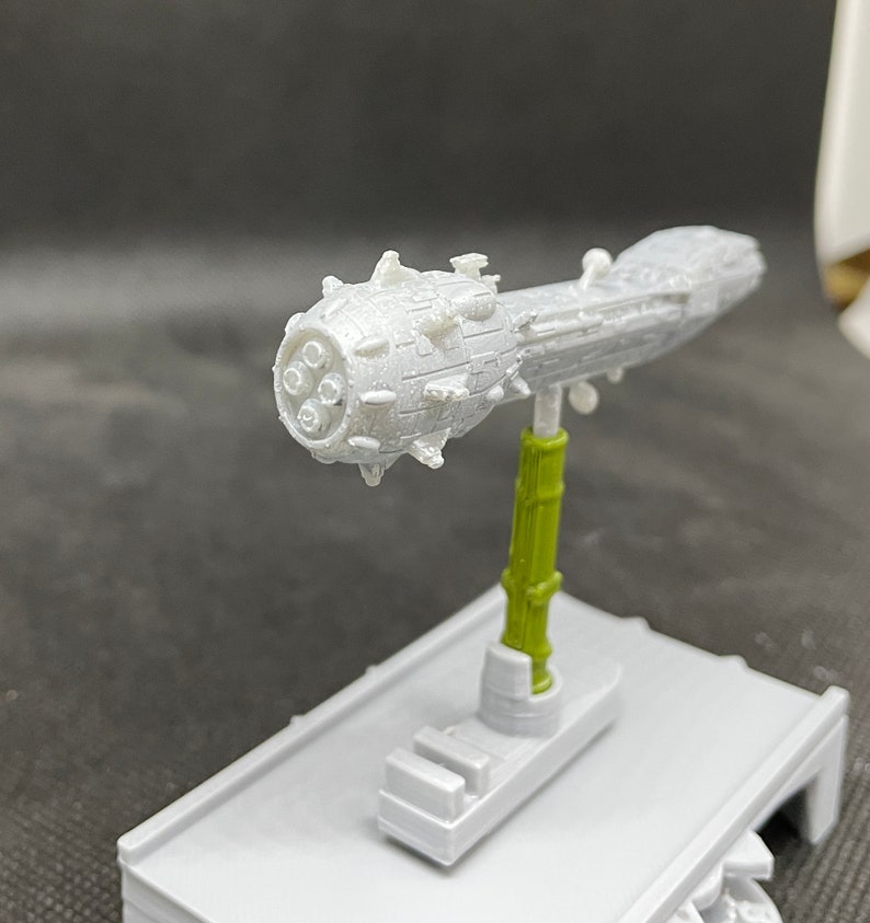 May include: A white 3D printed model of a spaceship with a spiky, round front section and a long, thin body. The model is mounted on a green stand.