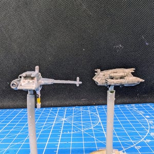 Op de afbeelding: Twee ongeverfde miniatuur-ruimteschepen op standaards. Het ruimteschip links is een grijs, lang, dun schip met een puntige neus en een lange, dunne staart. Het ruimteschip rechts is een grijs, kort, breed schip met een afgeronde neus en een korte, brede staart.