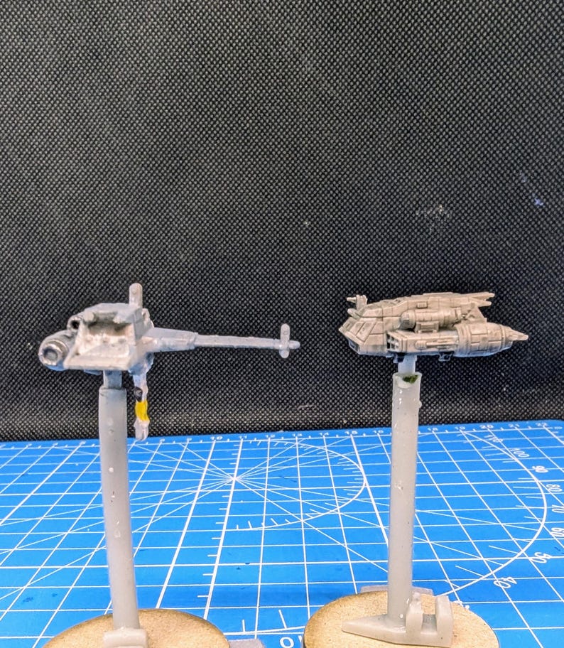 Op de afbeelding: Twee ongeverfde miniatuur-ruimteschepen op standaards. Het ruimteschip links is een grijs, lang, dun schip met een puntige neus en een lange, dunne staart. Het ruimteschip rechts is een grijs, kort, breed schip met een afgeronde neus en een korte, brede staart.