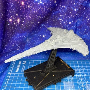 May include: A detailed, grey spaceship model on a black stand. The spaceship features intricate detailing and a unique wing design. The stand is labelled "MUNIFICENT". The background is a blue and purple galaxy design.