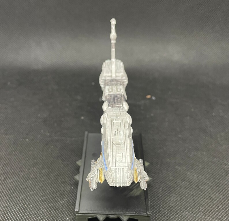 May include: A silver and blue model of a spaceship with a long, thin body and a pointed nose. The model is on a black base with a textured surface.