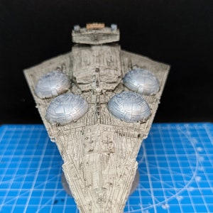 May include: A detailed model of a Star Destroyer spaceship, painted in a weathered gray color. The model features four large, round, silver-colored engine exhaust ports on the top of the ship. The model is sitting on a blue cutting mat.