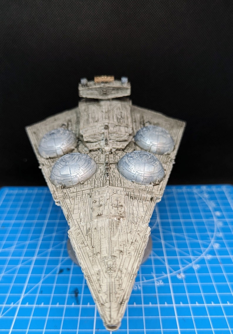 May include: A detailed model of a Star Destroyer spaceship, painted in a weathered gray color. The model features four large, round, silver-colored engine exhaust ports on the top of the ship. The model is sitting on a blue cutting mat.