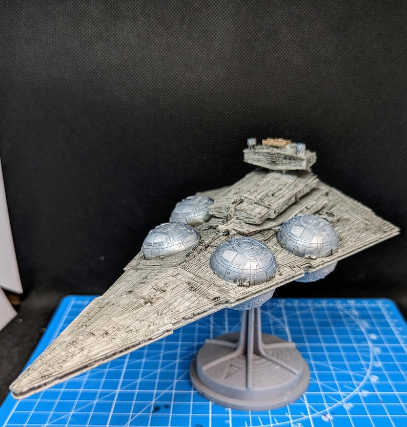 May include: A gray and white model of a Star Destroyer spaceship with a detailed design. The model is on a gray stand and has a textured surface.