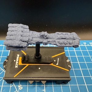 Op de afbeelding: Een grijs, gedetailleerd model van een ruimteschip, gemonteerd op een zwarte standaard met het woord "PINNACLE" in oranje. De basis heeft hoekige oranje accenten en een wijzerplaat met cijfers. Het model is zeer gedetailleerd.