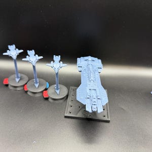 Puede incluir: Una colección de modelos de naves espaciales en miniatura de color azul claro. La imagen muestra tres naves más pequeñas sobre soportes con detalles rojos y azules, y una nave más grande sobre una base negra.