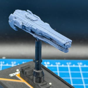 May include: A light blue, detailed model of a spaceship, mounted on a black stand. The spaceship has a long, rectangular body with intricate surface details, including what appear to be turrets and panels. The stand is attached to a black base with yellow accents.