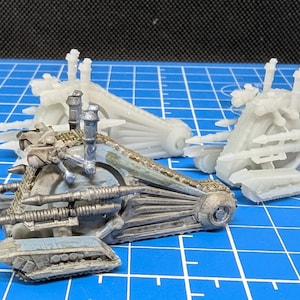 May include: A detailed model of a futuristic vehicle with a blue and silver paint scheme. The vehicle has a large, round wheel and multiple cannons on the front. The model is partially assembled and is sitting on a blue grid surface.