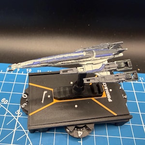 May include: A model spaceship with a blue and white design, mounted on a black base with orange accents. The base has the word "PINNACLE" printed on it. The model is set against a blue grid background.