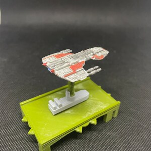May include: A white and red spaceship model with gray details, mounted on a green base. The spaceship has a long, narrow body with a pointed nose and wings. It has several small windows and a few small, round details on the body.