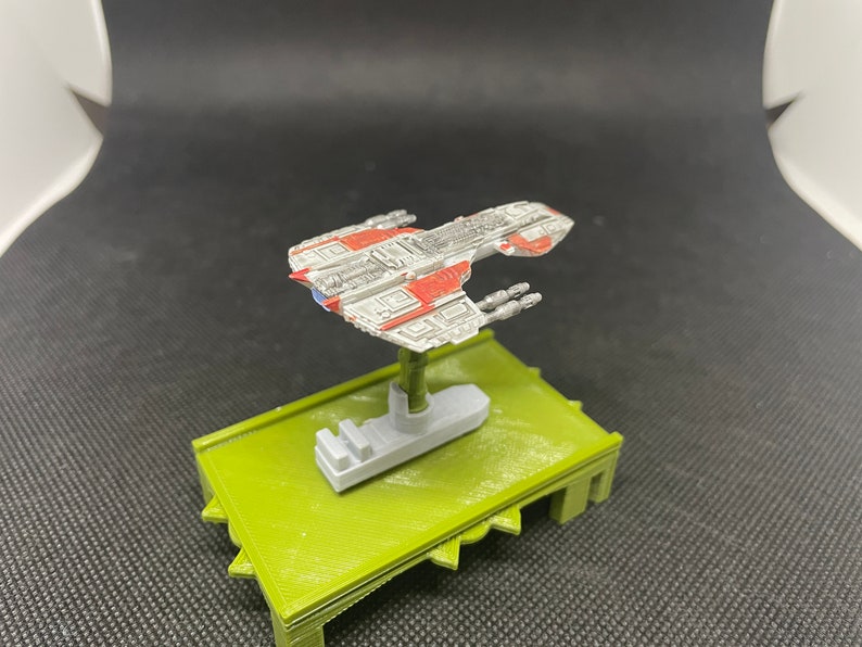 May include: A white and red spaceship model with gray details, mounted on a green base. The spaceship has a long, narrow body with a pointed nose and wings. It has several small windows and a few small, round details on the body.