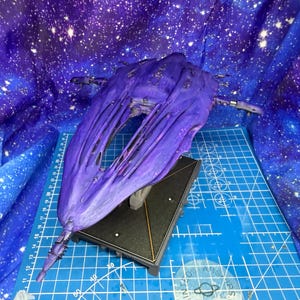 Könnte beinhalten: Ein lila Raumschiffmodell mit komplizierten Details, das auf einem schwarzen Ständer ruht. Das Modell hat eine strukturierte Oberfläche und befindet sich vor einem Hintergrund mit Sternengalaxie-Design. Das Modell ist etwa 30 cm lang.