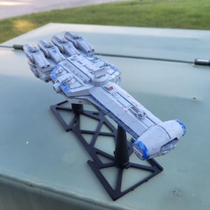 May include: A detailed scale model of a starship featuring a light gray hull with blue accents, multiple engine thrusters at the rear, and a long, slender forward section. The model is mounted on a black, geometric plastic display stand.
