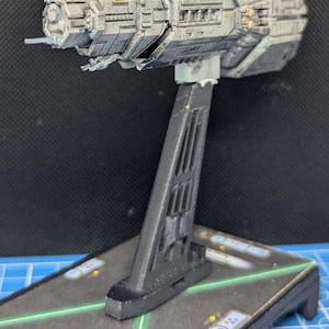May include: A gray and silver spaceship model on a black stand. The spaceship has a long, cylindrical body with a pointed nose and a series of windows and hatches along its side. The stand has a triangular base and a vertical support.