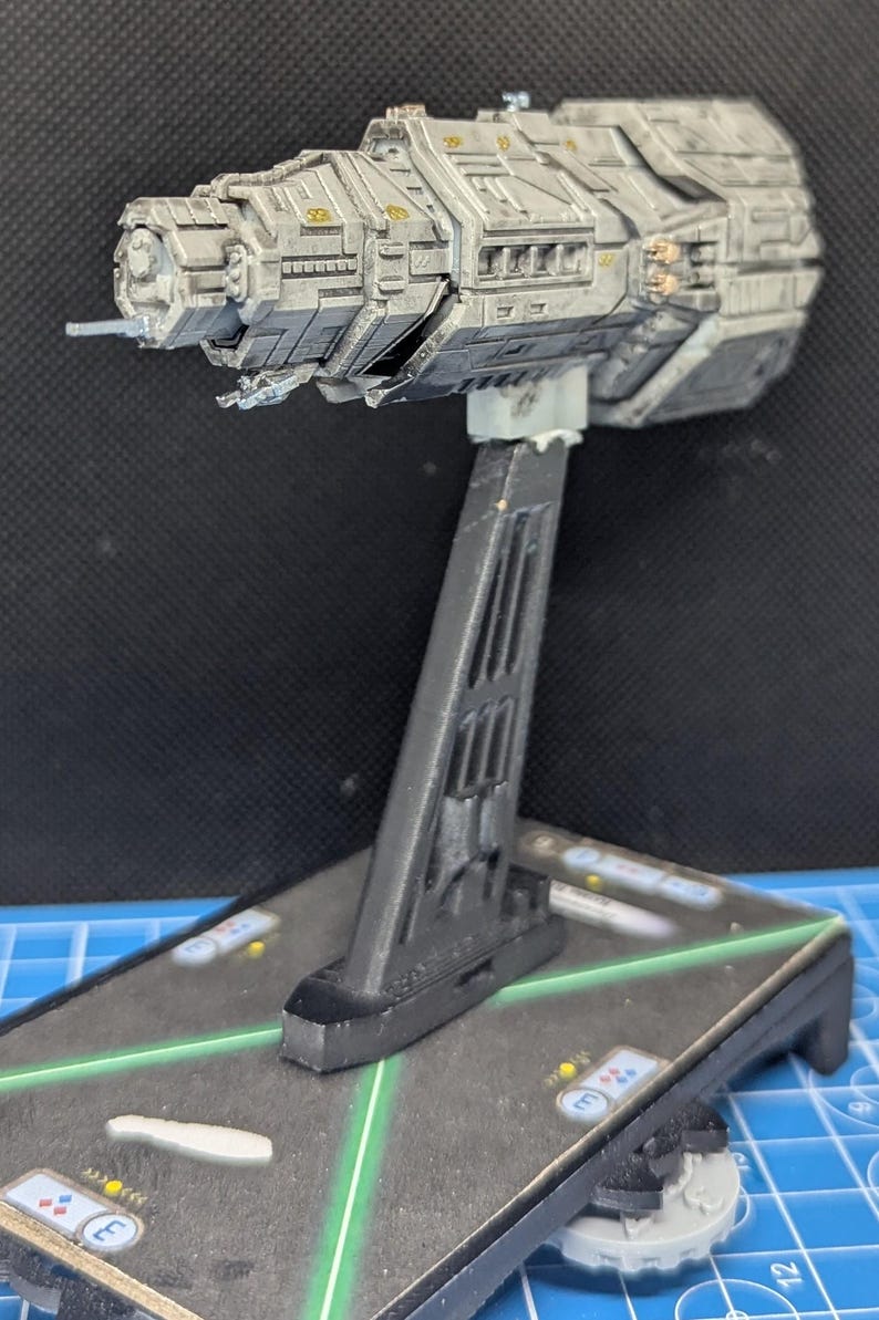 May include: A gray and silver spaceship model on a black stand. The spaceship has a long, cylindrical body with a pointed nose and a series of windows and hatches along its side. The stand has a triangular base and a vertical support.