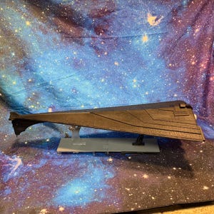 Puede incluir: Modelo gris oscuro de una nave espacial, con un diseño largo y elegante y detalles de superficie. El modelo está montado sobre una base gris claro con soportes transparentes, sobre un fondo de galaxia estrellada.