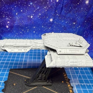 Peut inclure: Maquette de vaisseau spatial grise avec des panneaux détaillés et une conception à plusieurs niveaux. Le modèle est monté sur un support noir, avec le mot "MUNIFICENT" visible sur la base. Le fond est un ciel étoilé bleu.