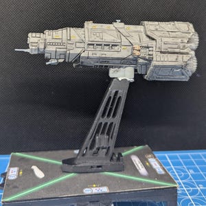 May include: A gray and black spaceship model with a detailed design, mounted on a black plastic stand. The model is on a green and black gaming mat.