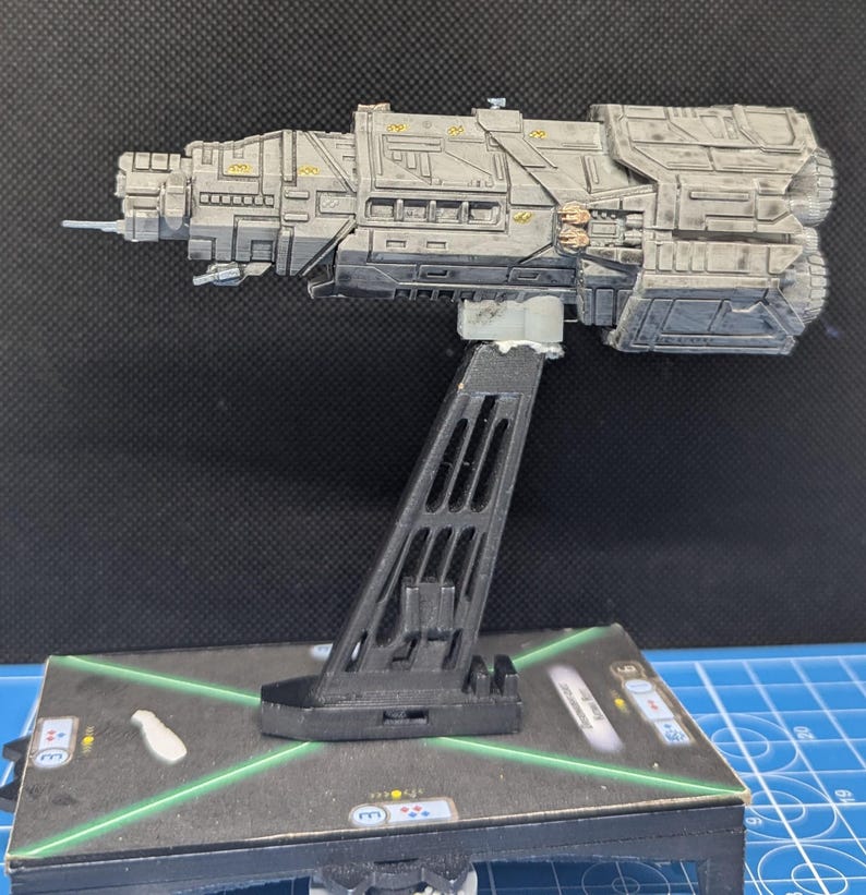 May include: A gray and black spaceship model with a detailed design, mounted on a black plastic stand. The model is on a green and black gaming mat.