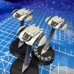 May include: Three miniature spacecraft models, predominantly white with black and grey detailing. Each model is mounted on a grey stand and a black base, with numbered markings. The background is a blue grid.