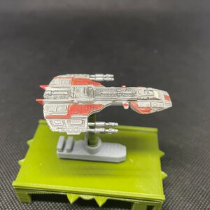 May include: A gray and red spaceship model with a detailed design. The model is mounted on a green base.