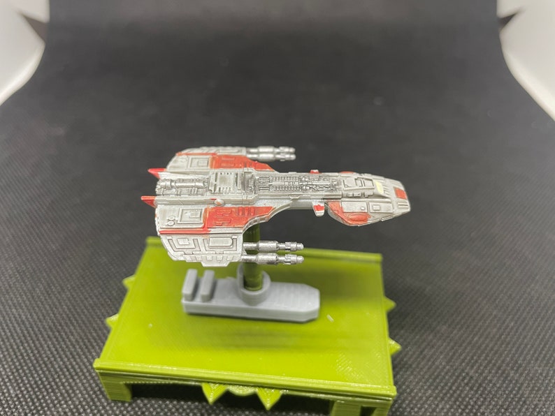 May include: A gray and red spaceship model with a detailed design. The model is mounted on a green base.