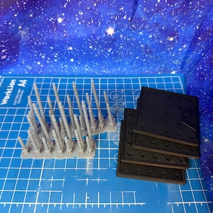 May include: A collection of gray plastic model parts, including multiple sprues of cylindrical components and several black square plates. The items are arranged on a blue cutting mat with grid lines, against a starry background.