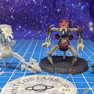 Puede incluir: Tres modelos de robots en miniatura. Uno es gris y sin montar. Otro es rojo y dorado, completamente montado sobre una base negra. Una tercera pieza roja está separada. El fondo es una cuadrícula azul, con un cielo estrellado.