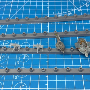 Könnte beinhalten: Vier graue Kunststoff-Aufbewahrungsregale mit einem Star Wars-Logo an jedem Ende. Die Regale haben jeweils 17 Löcher und dienen zur Aufbewahrung von kleinen Modell-Raumschiffen. Die Raumschiffe sind aus Kunststoff und in verschiedenen Farben lackiert, darunter Grau, Weiß und Blau.