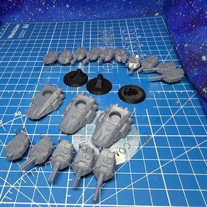 May include: A collection of unpainted, gray miniature spaceship models and bases. The models vary in size and design, with some featuring turrets and detailed paneling. The items are arranged on a blue grid cutting mat.